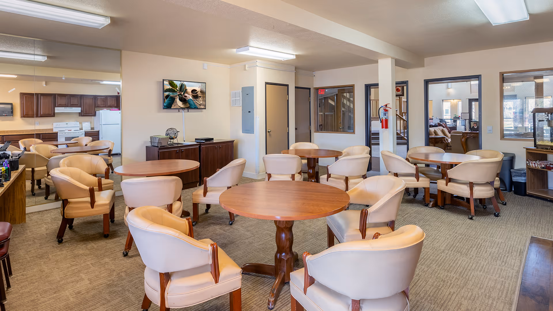 A bright common area with multiple round wooden tables surrounded by beige cushioned chairs. The room has beige walls and carpeted floors. In the background, there is a kitchen area with wooden cabinets, a white stove, and a refrigerator. A flat-screen TV is mounted on the wall above a wooden cabinet. There are windows and doors leading to other rooms, and a fire extinguisher is mounted on a pillar.