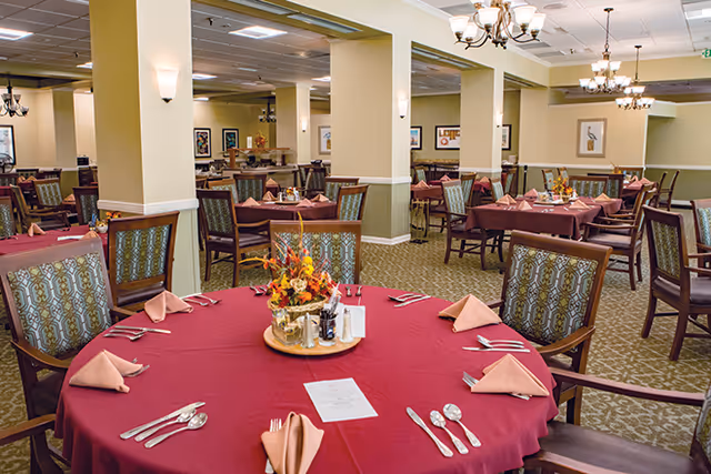 Spacious dining room with round tables set with burgundy tablecloths, folded napkins, floral centerpieces and upholstered chairs under chandeliers.