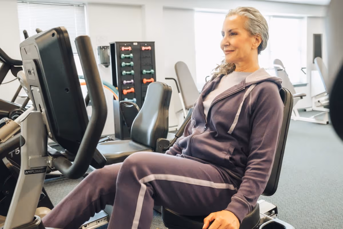 An older woman in a purple tracksuit is seated on a recumbent exercise bike in a fitness room with various exercise equipment and colorful dumbbells in the background.