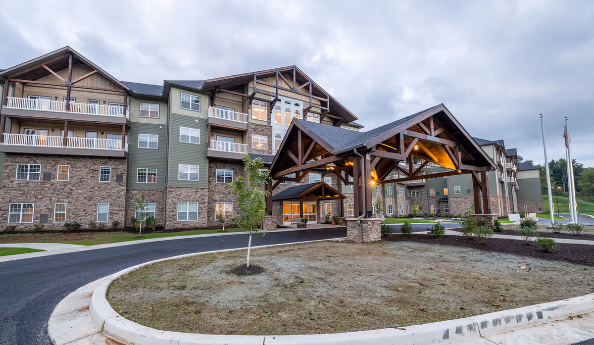 Exterior view of a large senior living facility building with stone and green siding, multiple balconies, and a covered entrance with wooden beams and lighting. The driveway curves around a small landscaped area with young trees and shrubs under a cloudy sky.