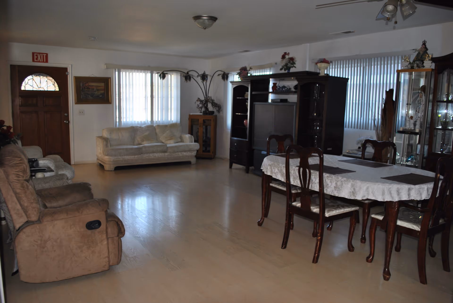 A spacious living room with light wood flooring, featuring a brown recliner chair, a beige sofa, a wooden dining table with six chairs covered with a white tablecloth, and a large dark wood entertainment center with a TV. The room has large windows with vertical blinds, a wooden front door with a glass window, and various decorative items including flowers and figurines on shelves and cabinets.