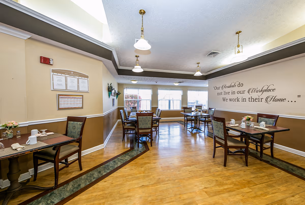 A bright dining room with wooden floors and several tables set with placemats, napkins, cups, and small flower arrangements. The walls are painted beige and brown, with a quote on one wall that reads, 'Our Residents do not live in our Workplace We work in their Home...'. Large windows allow natural light to fill the room, and pendant lights hang from the ceiling.