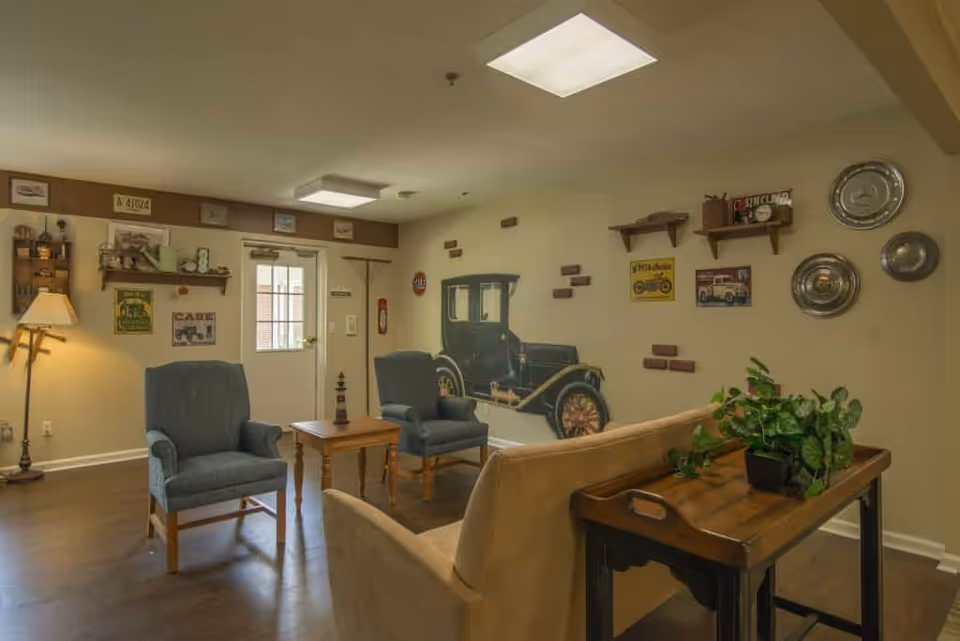 Cozy common room with armchairs and a couch, wooden tables, plants, and a vintage car mural on the wall.