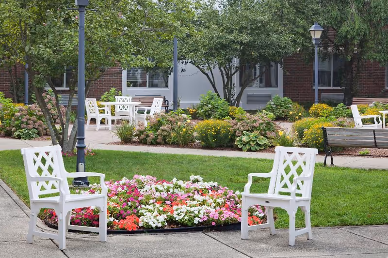 Outdoor courtyard with white chairs around a colorful flower bed in front of a brick building.