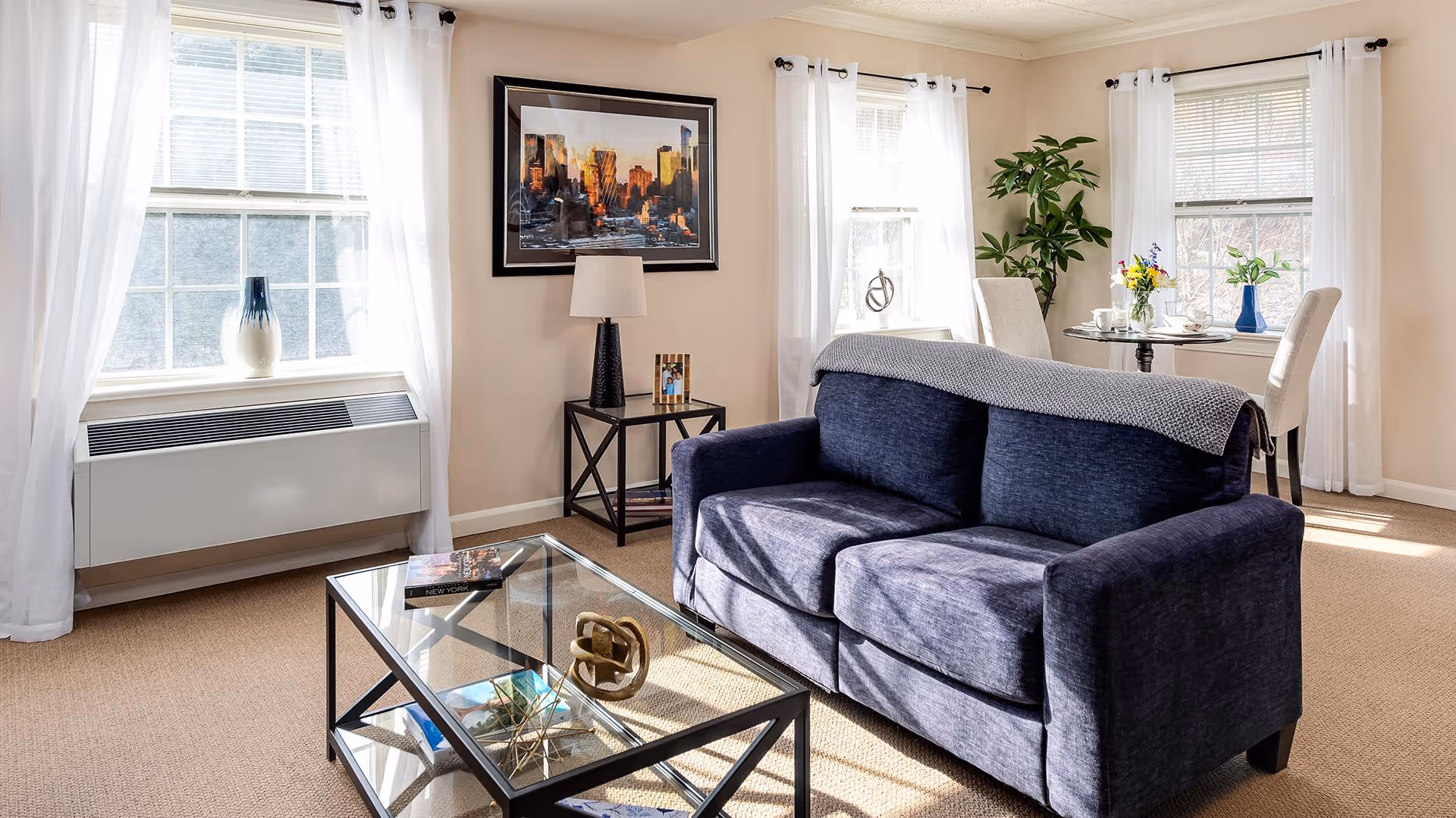 Well-lit living room featuring a navy sofa, glass coffee table, side table with a lamp and artwork, and a small dining table by the windows.