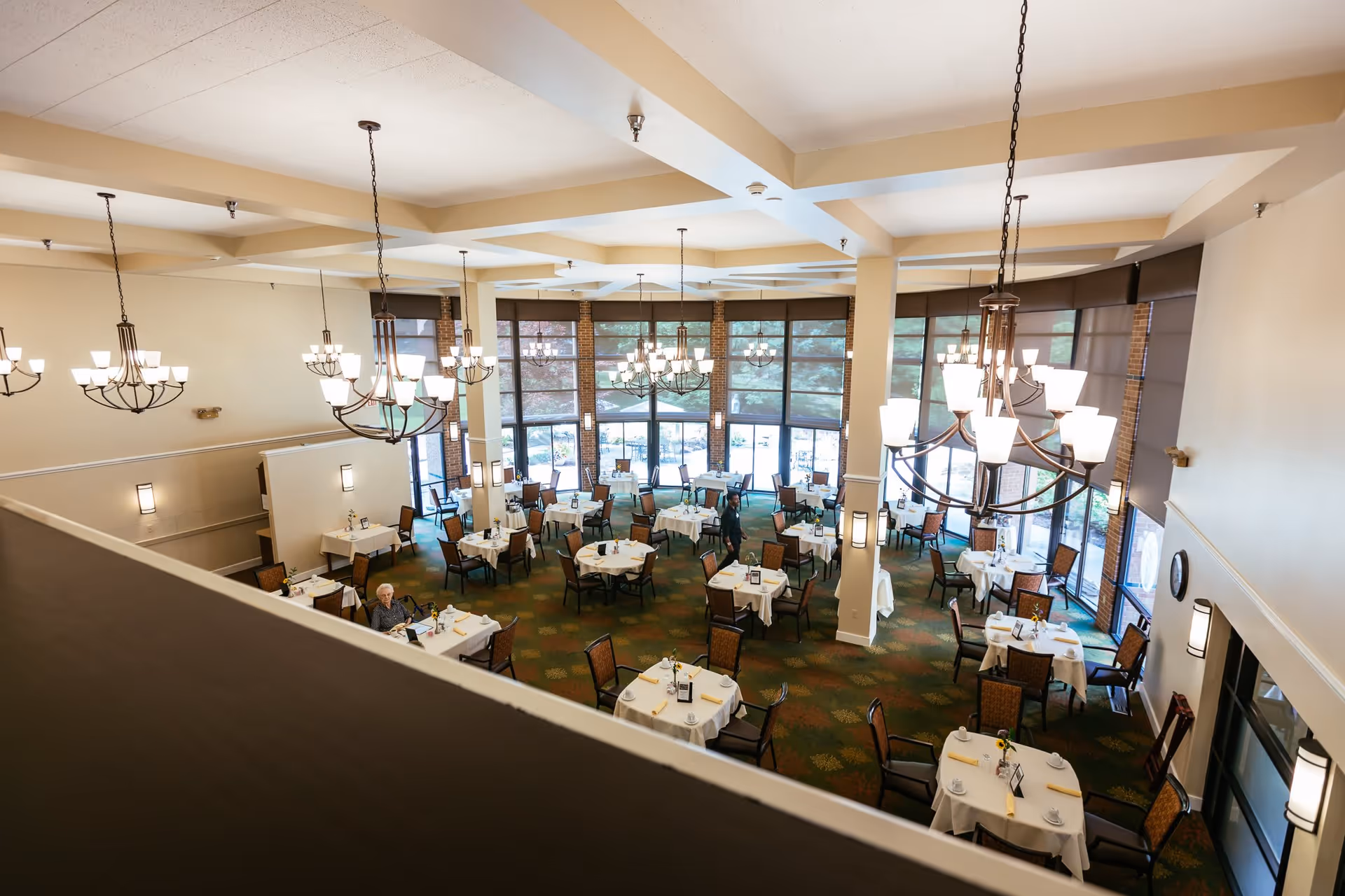A spacious dining room with multiple round tables covered with white tablecloths, each set with cups, napkins, and small flower arrangements. The room has large windows with brown blinds, green carpet with a leaf pattern, and several chandeliers hanging from the ceiling. Two people are visible, one seated at a table and another standing near the center of the room.