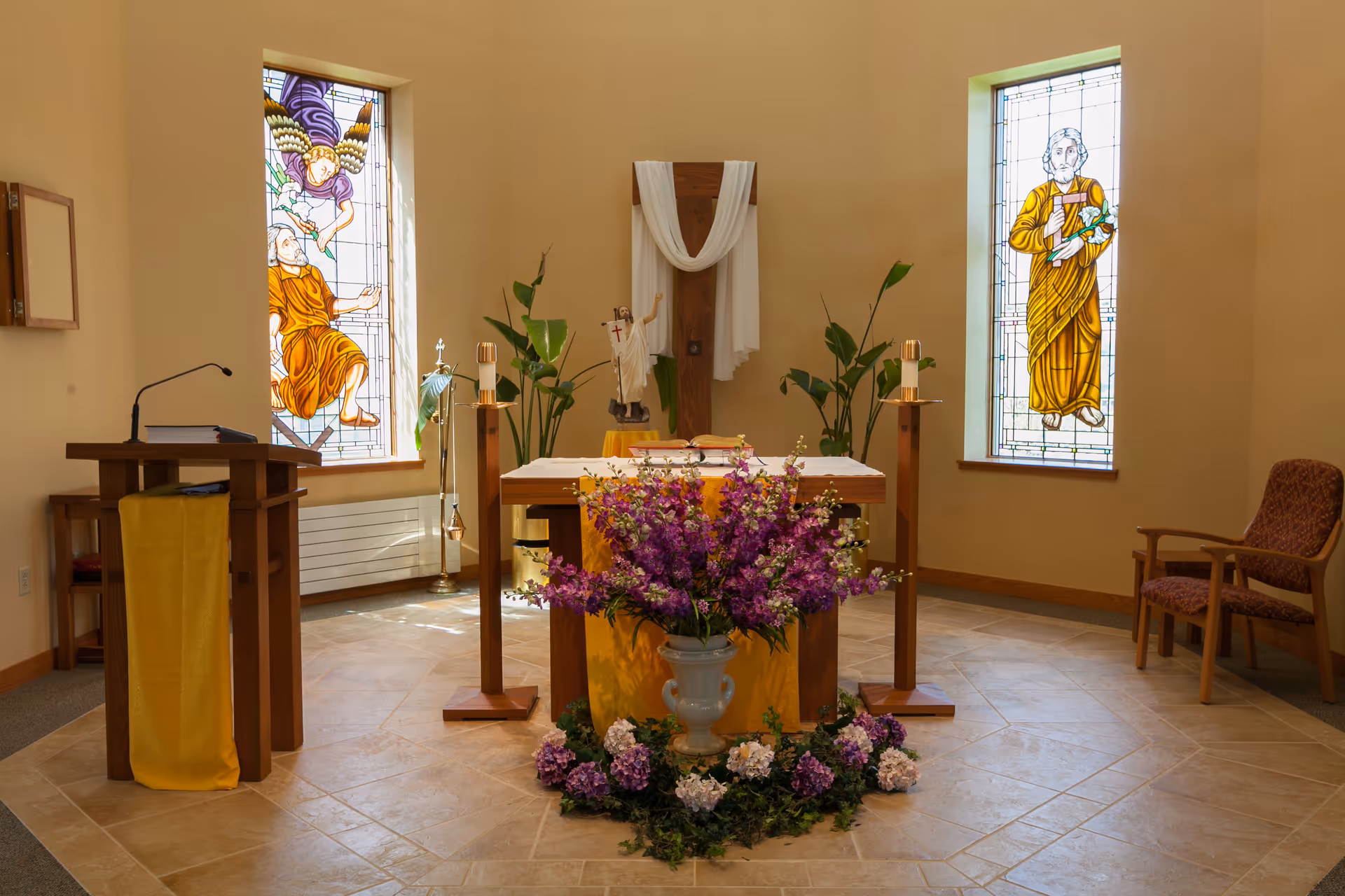 Interior view of a small chapel with a wooden altar adorned with purple and white flowers. Behind the altar is a wooden cross draped with white cloth, flanked by two tall candles. There are two stained glass windows depicting religious figures on either side of the altar. A wooden lectern with a yellow cloth is on the left, and a single upholstered chair is on the right.