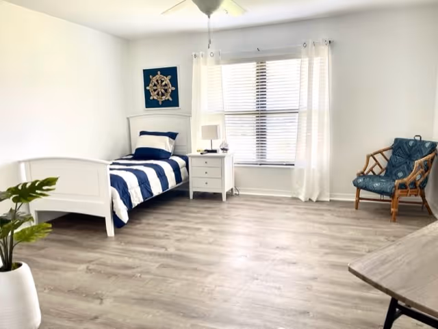 A bright and spacious bedroom with a single white bed featuring navy blue and white striped bedding. Next to the bed is a white nightstand with a lamp and decorative items. A large window with white curtains allows natural light to fill the room. There is a blue cushioned chair with a wooden frame in the corner and a potted plant in a white vase on the floor. The room has light wood flooring and white walls.