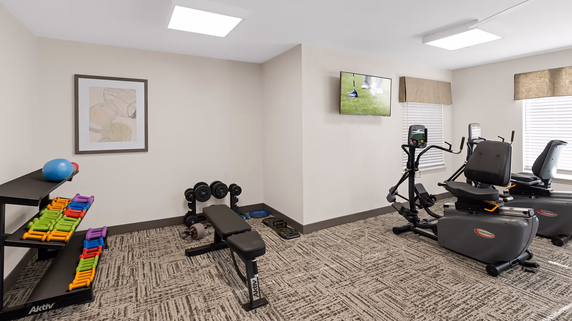 A fitness room with exercise equipment including a weight bench with dumbbells, colorful hand weights on a rack, two recumbent exercise bikes, and a wall-mounted TV showing a golf game. The room has beige walls, patterned carpet, and two windows with blinds and valances.