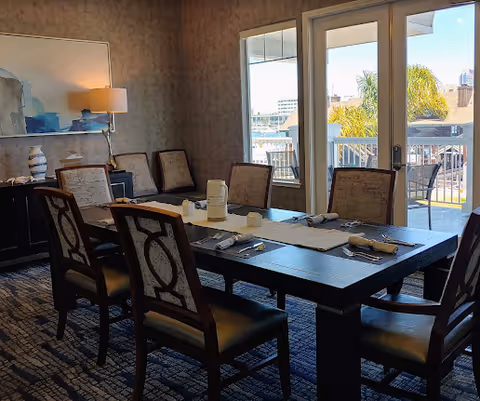 A dining room with a dark wooden table set for six people with placemats, napkins, and utensils. There are six upholstered chairs around the table. A sideboard with decorative items and a lamp is against the wall. Large glass doors open to an outdoor patio area with chairs and a view of buildings and palm trees outside.