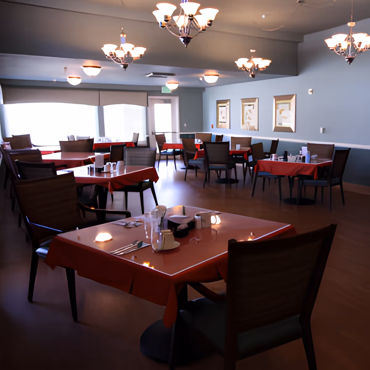 Dining room with red-clothed tables, chairs, chandeliers, and place settings in a communal assisted-living dining area.