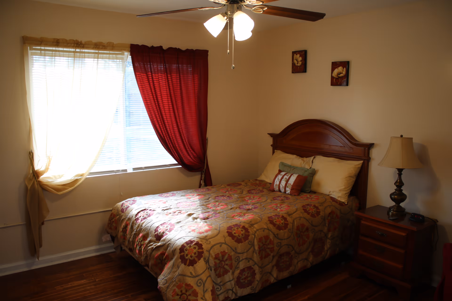 A cozy bedroom with a wooden bed frame and a floral patterned bedspread. The bed has three pillows, and there is a wooden nightstand with a lamp and a remote control. The room has a window with sheer beige and red curtains, and a ceiling fan with lights is visible above.