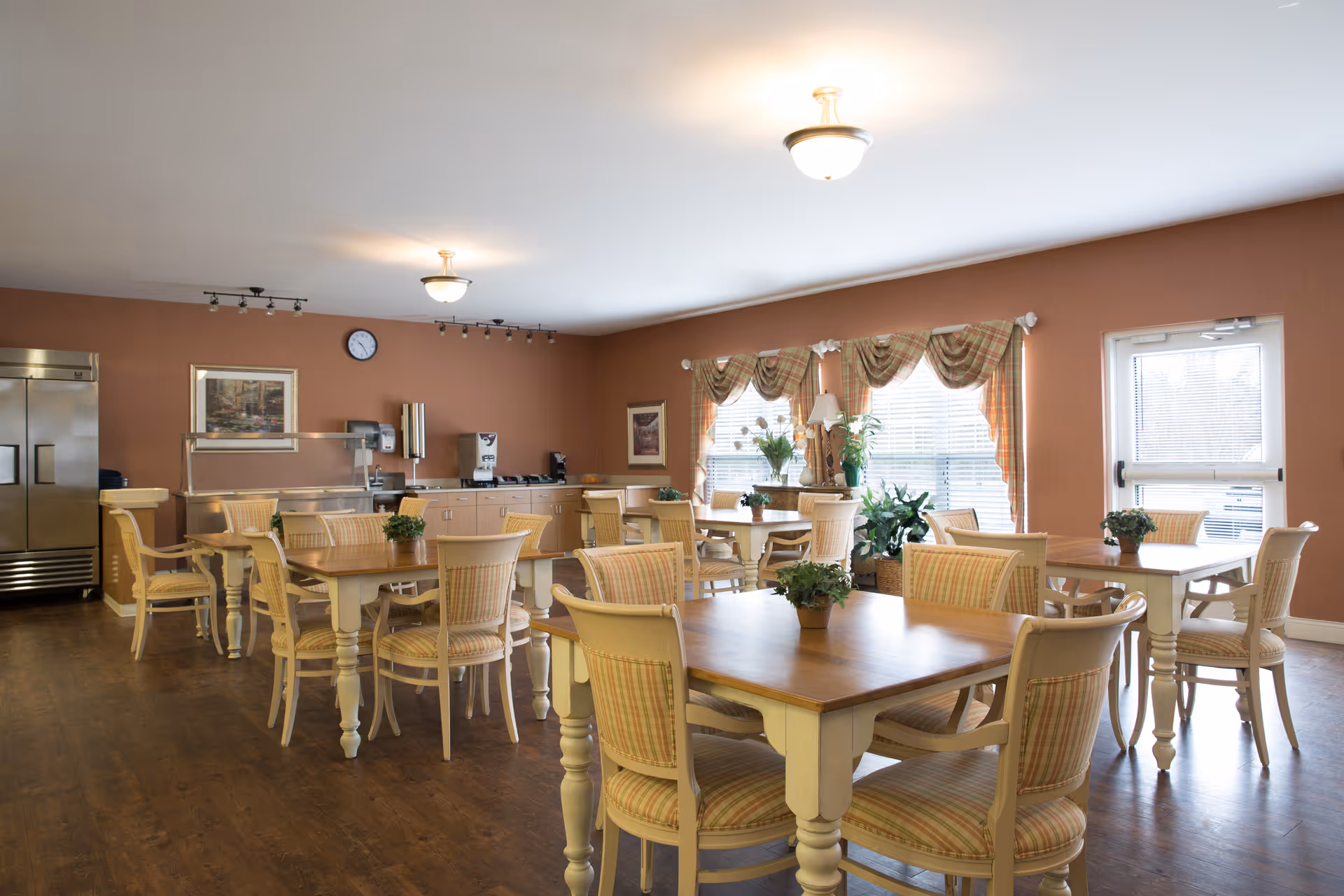 A bright dining room with several wooden tables and chairs arranged neatly. The chairs have striped cushions, and small potted plants are placed on each table. The room has large windows with plaid valances, letting in natural light. There is a stainless steel refrigerator and a counter with a coffee machine and other appliances along one wall. The walls are painted a warm brown color, and the floor is wooden.