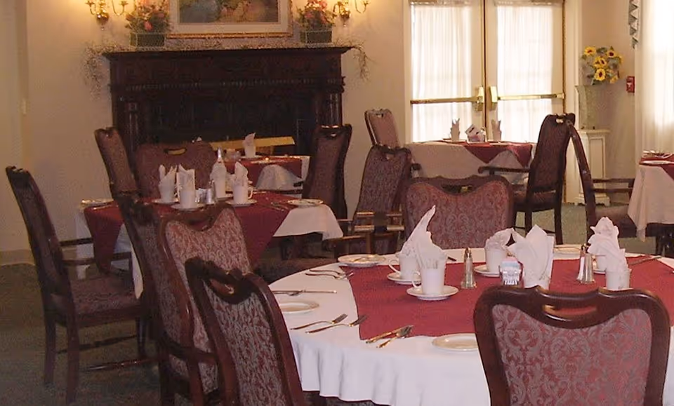 Dining room with round tables covered in white and burgundy tablecloths, set with white napkins, cups, plates, and silverware. Upholstered chairs surround the tables. A fireplace with floral decorations and wall sconces is visible in the background, along with large windows letting in natural light.