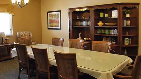 A dining room with a long table, chairs, and a wooden bookshelf.