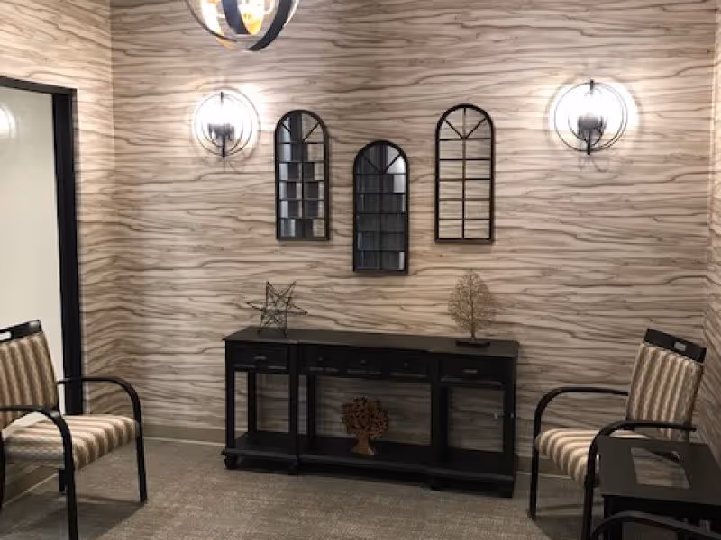 A small seating area with two striped armchairs facing each other, a black console table with decorative items including a star sculpture and a tree sculpture, three arched mirrors on a textured beige wall, and two wall-mounted light fixtures on either side of the mirrors.