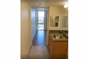 View down a narrow apartment interior hallway with a sink and countertop on the right leading to a windowed living area.