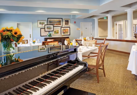 Interior view of a dining room in a senior living facility featuring a grand piano in the foreground, tables set with white tablecloths, napkins, and glassware, floral arrangements, framed artwork on the walls, and warm lighting.