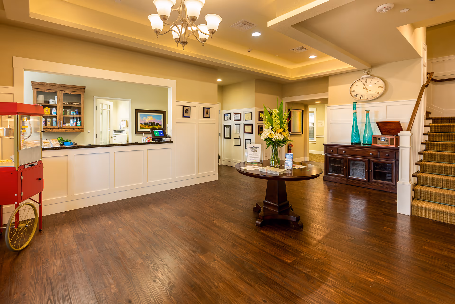 A bright and welcoming interior space with wooden flooring, a round wooden table with a flower arrangement in the center, a large clock on the wall above a wooden cabinet with decorative blue glass bottles, a staircase with carpeted steps, and a counter area with a popcorn machine on the left side.