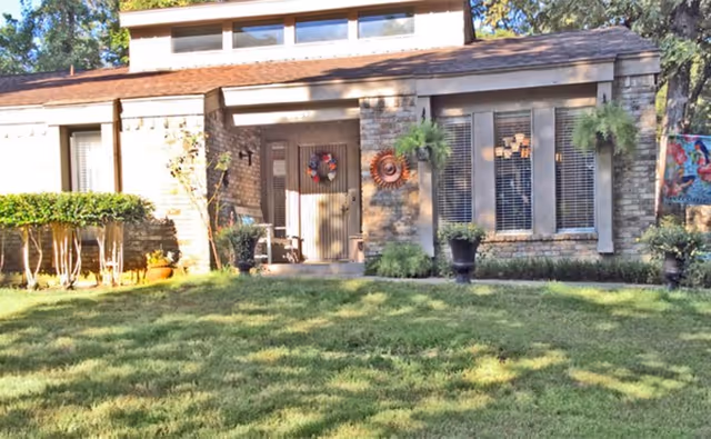 Front exterior view of a single-story building with a brick facade, a covered porch decorated with a wreath on the door, potted plants, and a well-maintained lawn in the foreground.