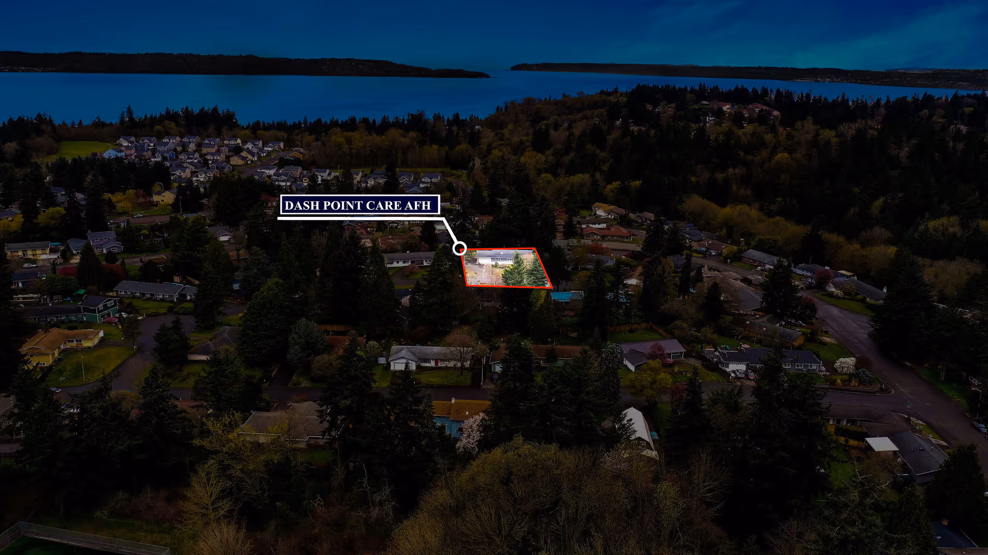 Aerial view of a residential neighborhood surrounded by dense trees and greenery, with a body of water visible in the background. A specific property is highlighted with a red outline and labeled 'DASH POINT CARE AFH'.