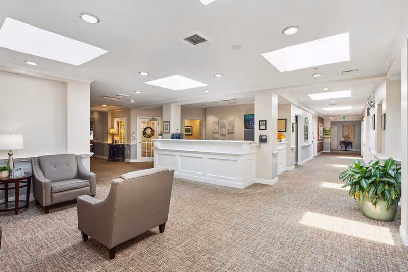 Bright and spacious senior living facility reception area with a white front desk, two gray armchairs, a side table with a lamp, potted plants, and artwork on the walls. The space is well-lit with recessed lighting and skylights, and has a carpeted floor.