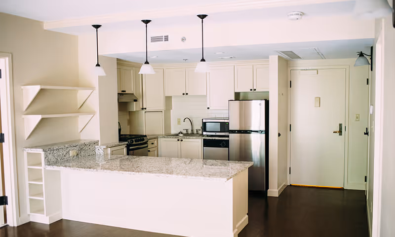 A modern kitchen area in an assisted living facility featuring white cabinetry, a granite countertop island with built-in shelving, stainless steel appliances including a refrigerator and microwave, a stove, and three pendant lights hanging from the ceiling. The kitchen is adjacent to a hallway with a closed door.