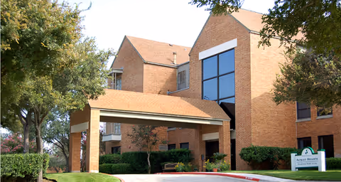 Exterior view of a brick building with a covered entrance and large windows, surrounded by trees and shrubs. A sign near the entrance reads 'Patriot Heights Assisted Living'.