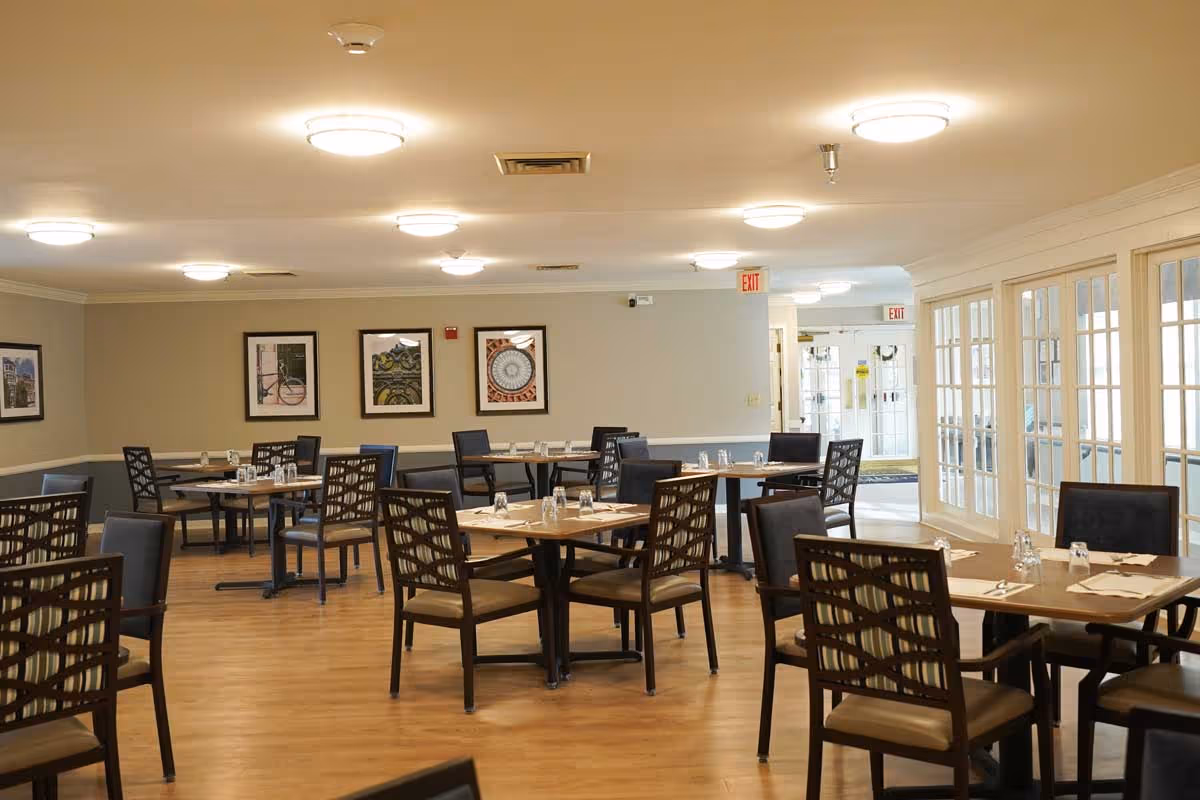 A dining room with multiple wooden tables and chairs arranged neatly. Each table is set with glasses and napkins. The room has wooden flooring, beige walls with framed artwork, and ceiling lights. There are glass-paneled doors on the right side leading to another area.