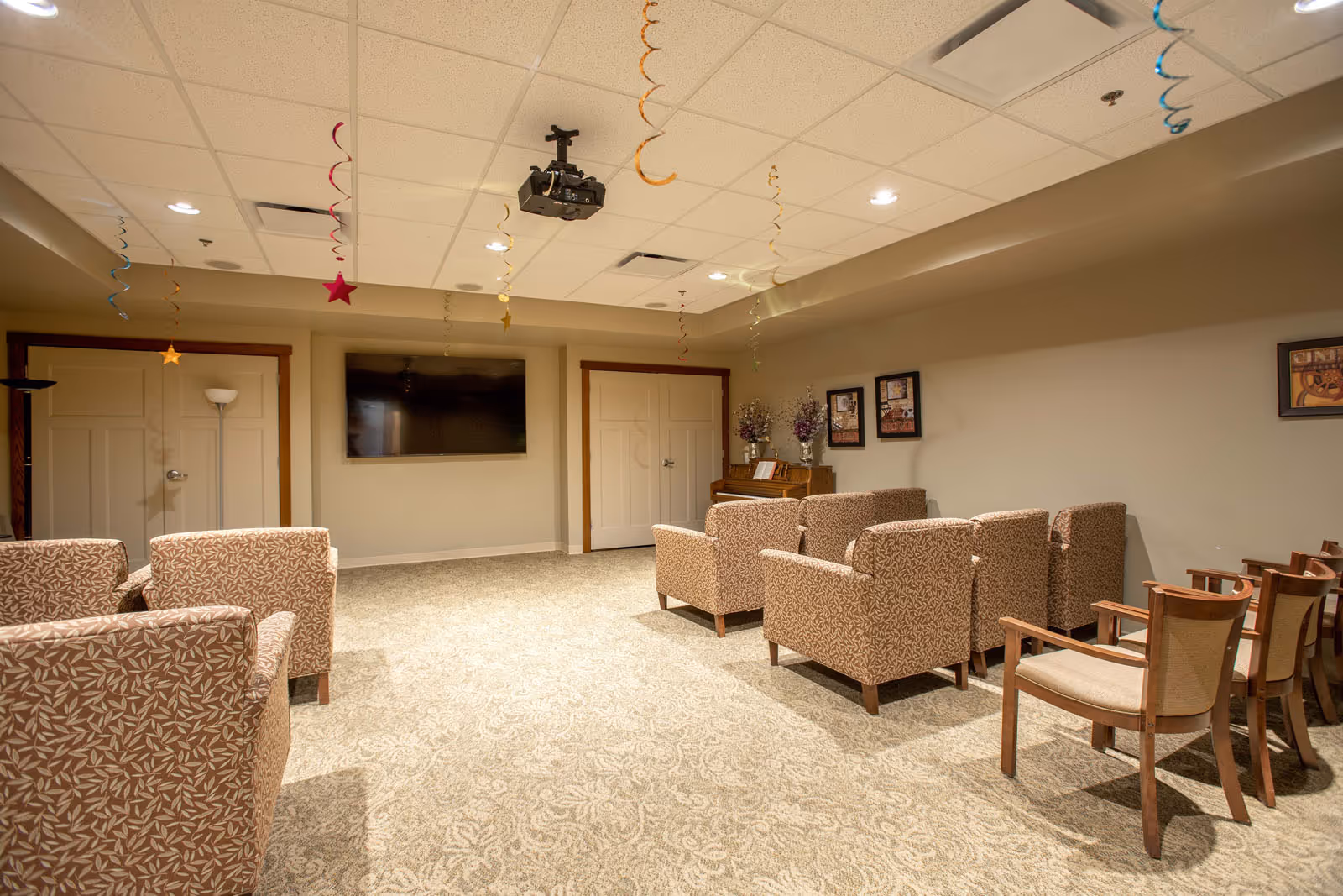 A cozy common room with patterned armchairs and wooden chairs arranged facing a wall-mounted flat screen TV. The ceiling has hanging colorful star decorations, and there is a piano with flower vases and framed pictures on the wall. The room has beige walls and carpeted floor with a subtle pattern.
