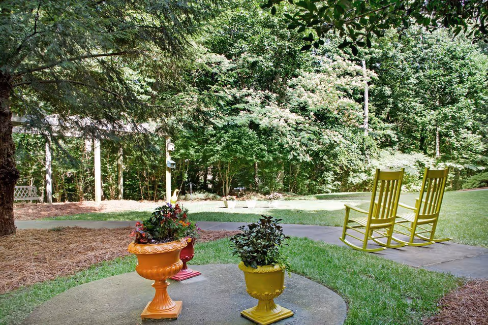 Two yellow rocking chairs on a paved patio facing a grassy yard and wooded area, with colorful potted plants in the foreground.