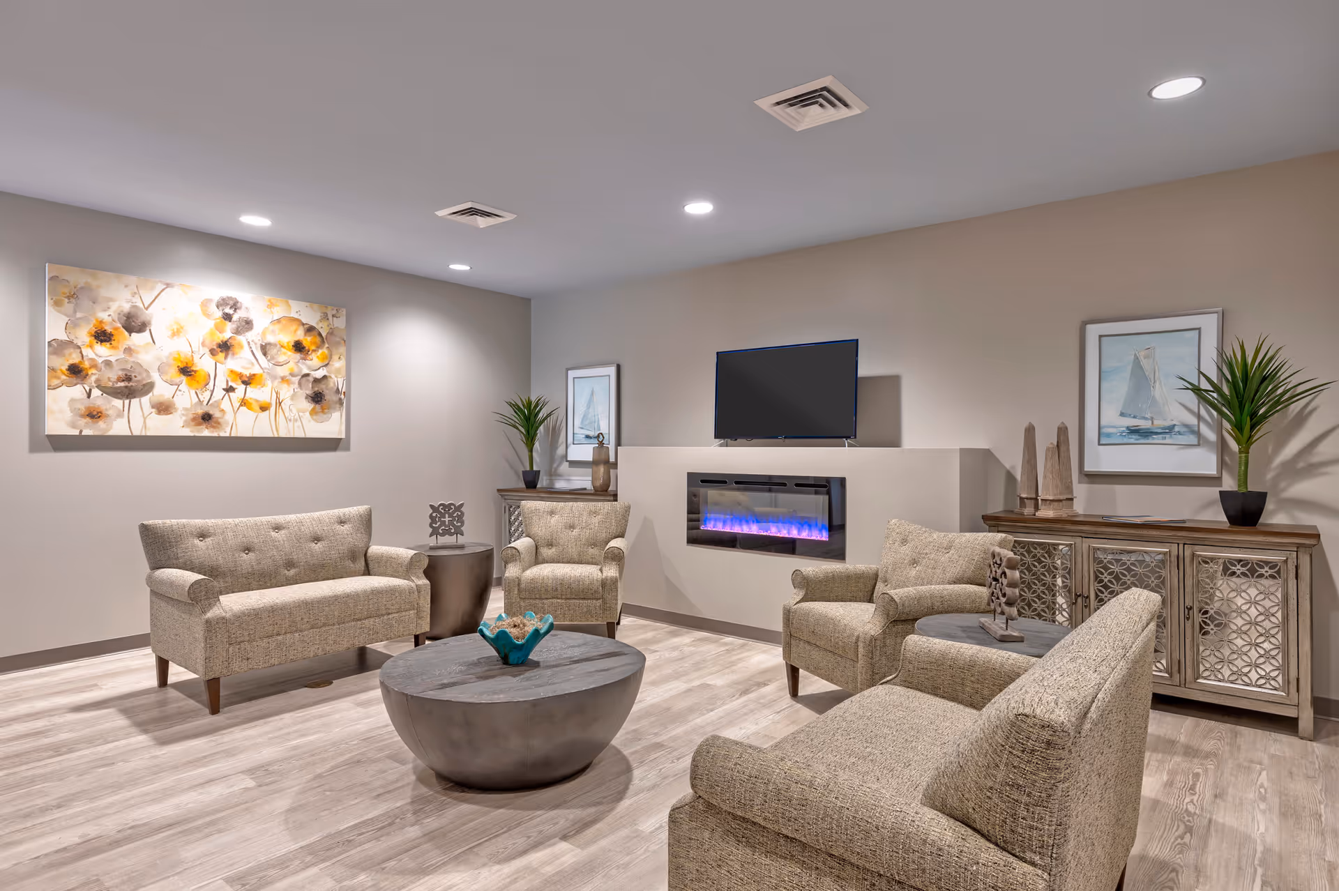 A modern lounge with upholstered sofas and armchairs arranged around a round coffee table, a wall-mounted TV above an electric fireplace, and decorative artwork and plants.
