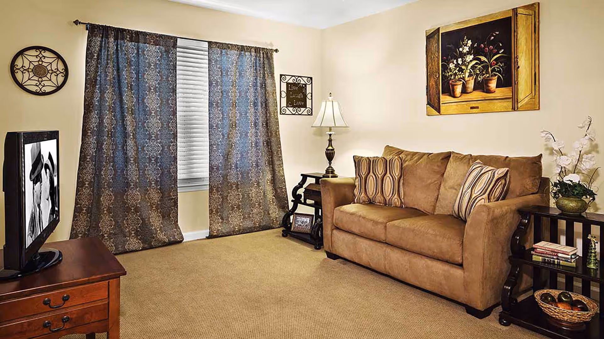 A cozy living room with a brown sofa adorned with patterned cushions, a wooden side table with a lamp, a small shelf with books and a decorative basket, a window with patterned curtains, and a wall painting of potted plants.