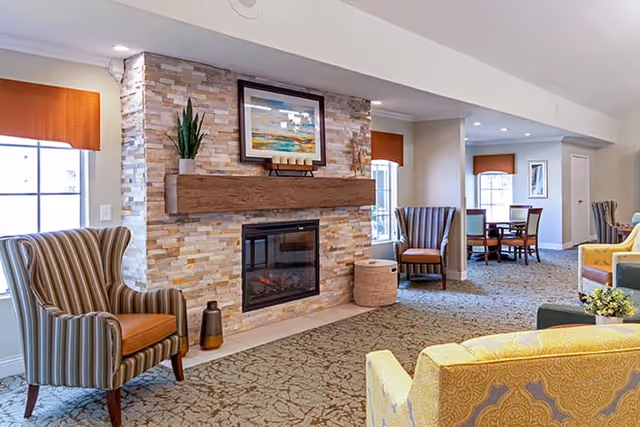 Cozy common room with a stone fireplace, wooden mantel and framed art, surrounded by upholstered armchairs and a carpeted seating area.