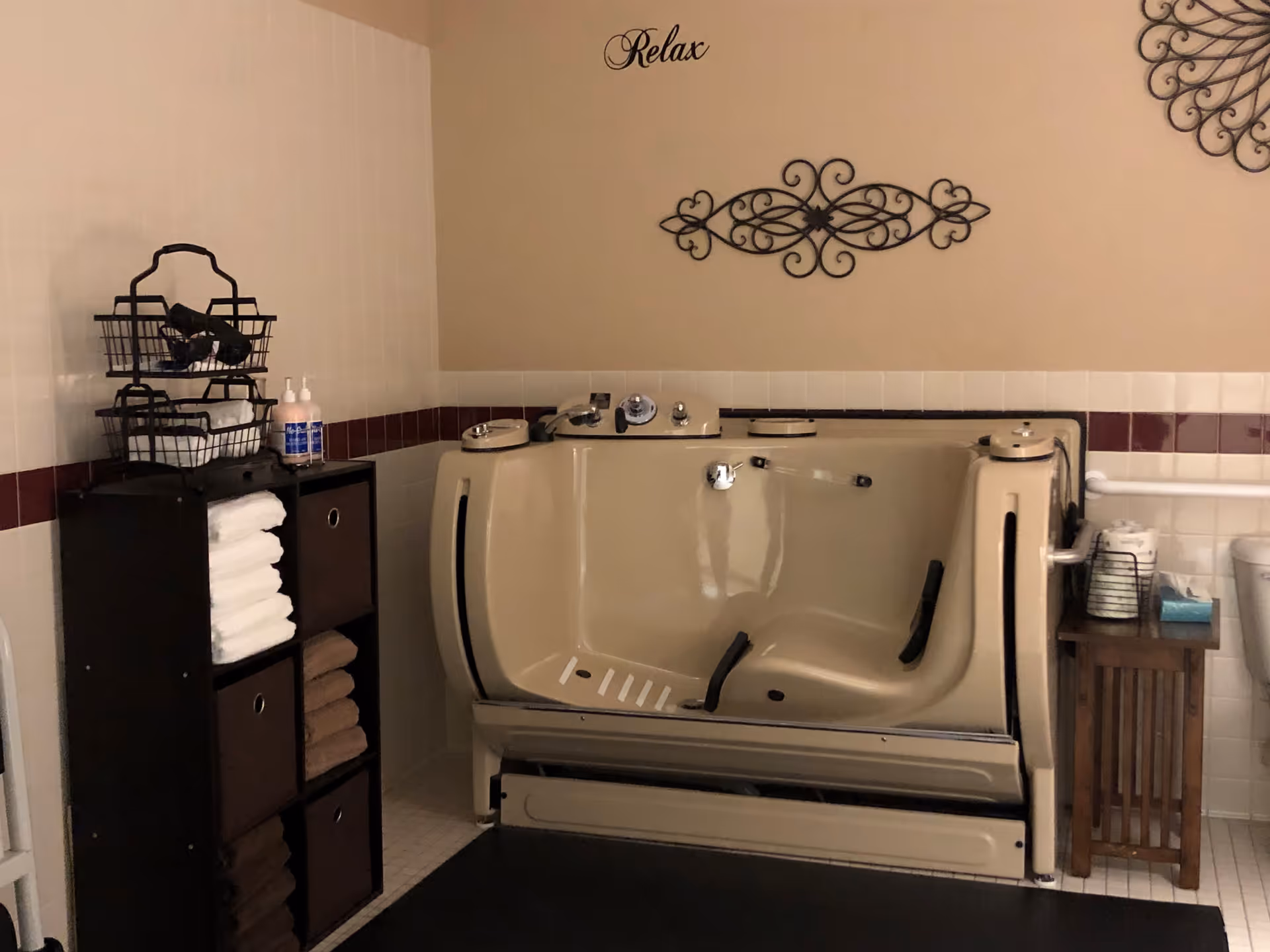 A beige walk-in bathtub with black handles in a bathroom setting. To the left is a black shelving unit holding neatly folded white and brown towels and two bottles of soap or lotion on top. On the right side, there is a small wooden table with a wire basket holding toilet paper and a box of tissues. The walls are tiled halfway up with white tiles and a maroon accent strip. Above the bathtub, the word 'Relax' is written on the beige wall along with decorative black metal wall art.