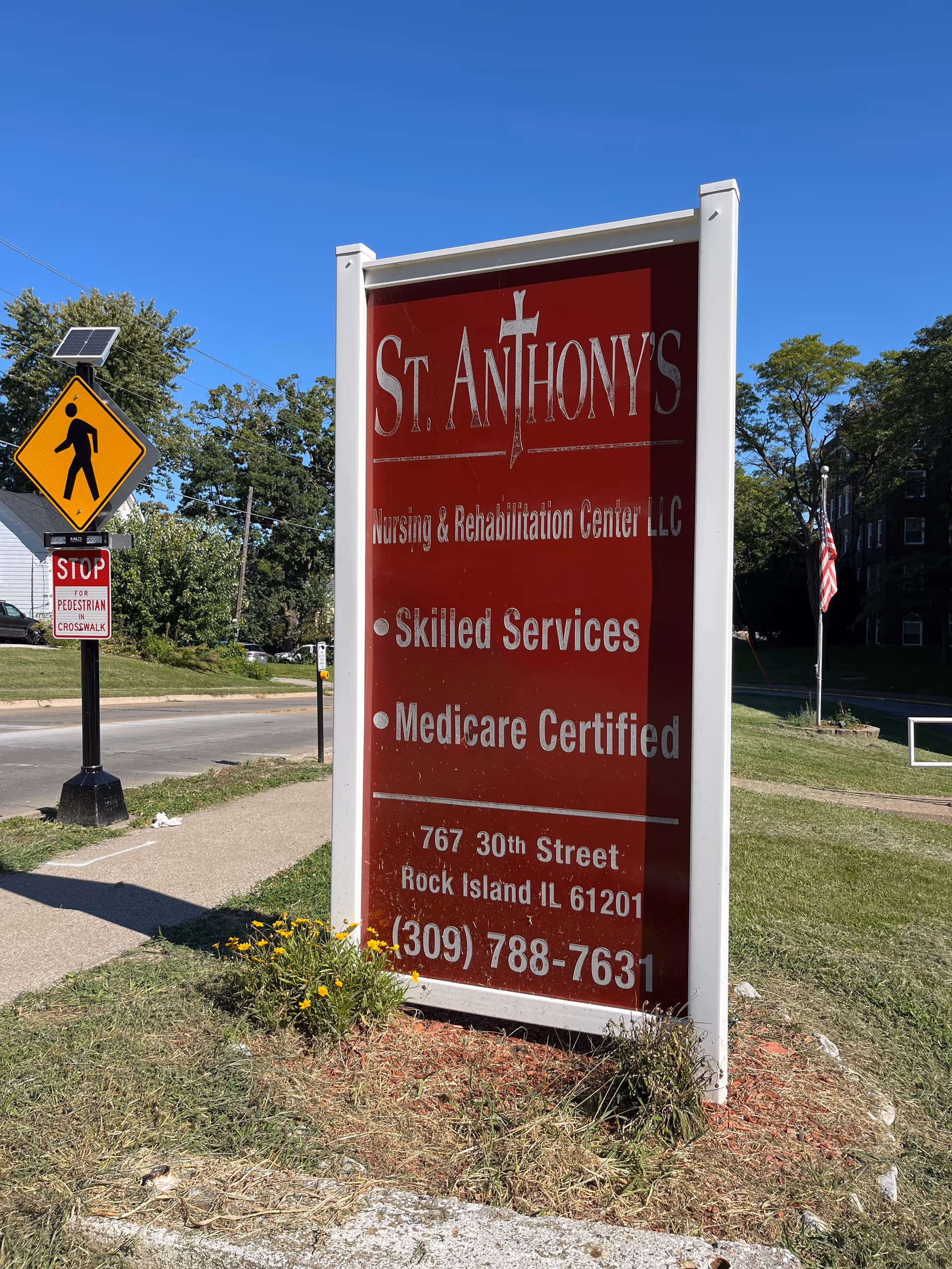 Outdoor image of a large red and white sign for St. Anthony's Nursing & Rehabilitation Center LLC, located on a grassy area near a sidewalk and street. The sign lists services including Skilled Services and Medicare Certified, along with the address 767 30th Street, Rock Island, IL 61201 and phone number (309) 788-7631. Nearby is a pedestrian crossing sign and a stop sign for pedestrian crosswalk. Trees and a building with an American flag are visible in the background under a clear blue sky.