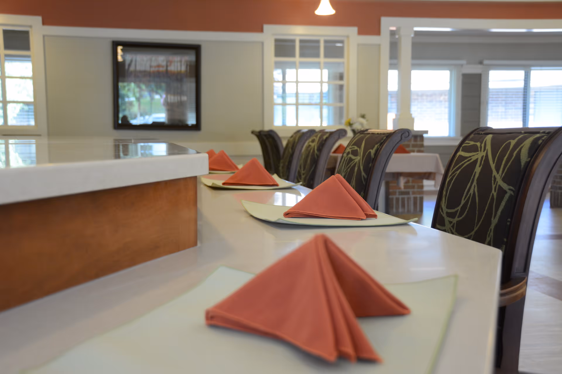 A dining area with a long white countertop set with folded red napkins on placemats. Upholstered chairs with a green and brown pattern are arranged along the counter. The room has large windows and a framed picture on the wall.