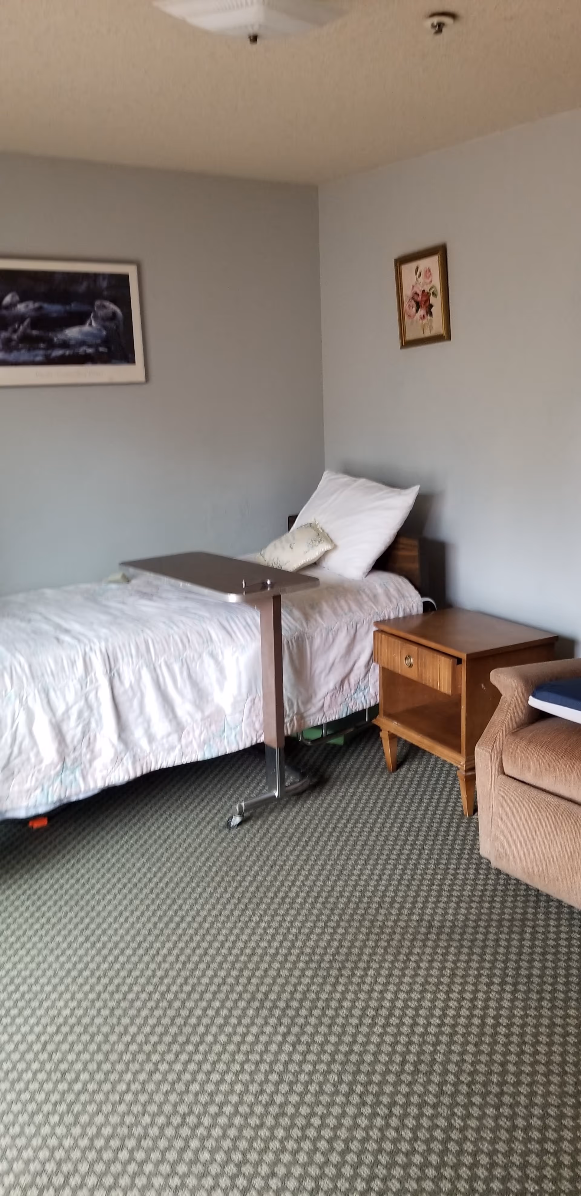 A simple bedroom with a single bed covered in a white quilt, a pillow, and a small cushion. There is a wooden nightstand next to the bed and a beige armchair partially visible on the right. Two framed pictures hang on the light gray walls. The floor is carpeted with a patterned design.