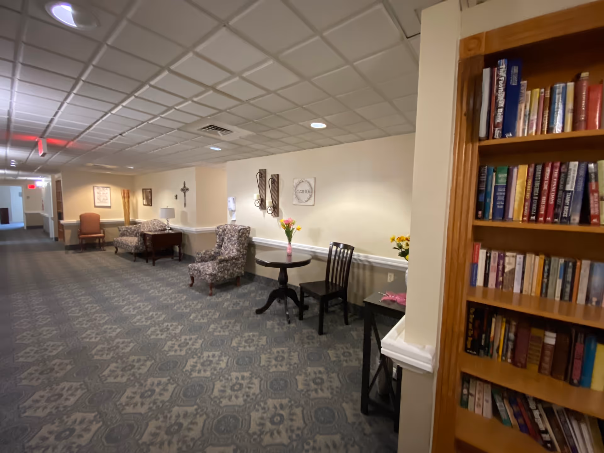 A hallway in a senior living facility with patterned carpet and a drop ceiling. Along the right wall, there is a small round table with a vase of flowers and a black chair. Further down the hallway, there are two upholstered armchairs and a small table with a lamp. On the left side, there is a wooden bookshelf filled with books. The walls are decorated with framed pictures and wall sconces.