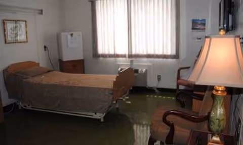 A senior living facility bedroom with a bed, chairs, and a window with blinds