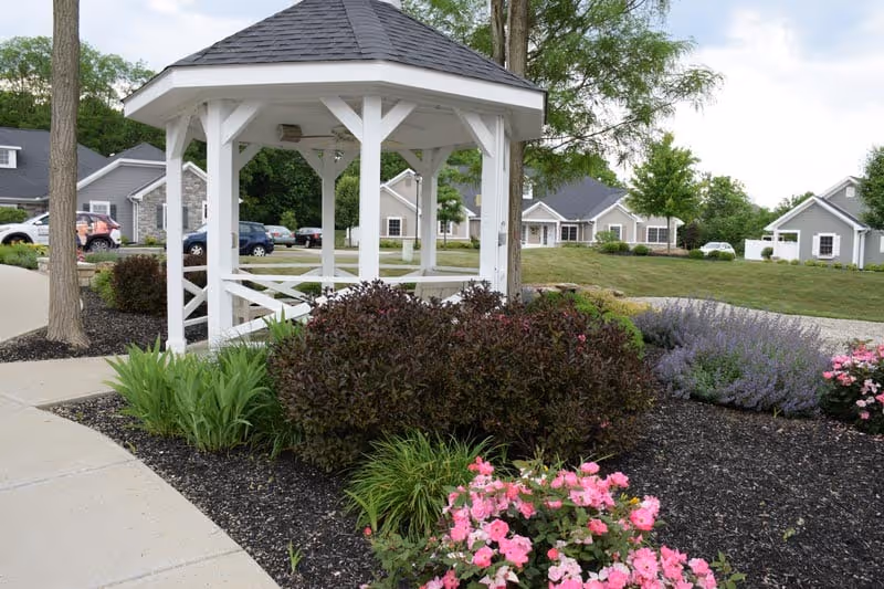 A white wooden gazebo with a gray shingled roof is situated in a landscaped garden area with various green shrubs and pink flowers. In the background, there are several single-story residential buildings with gray siding and white trim, along with parked cars and trees under a partly cloudy sky.