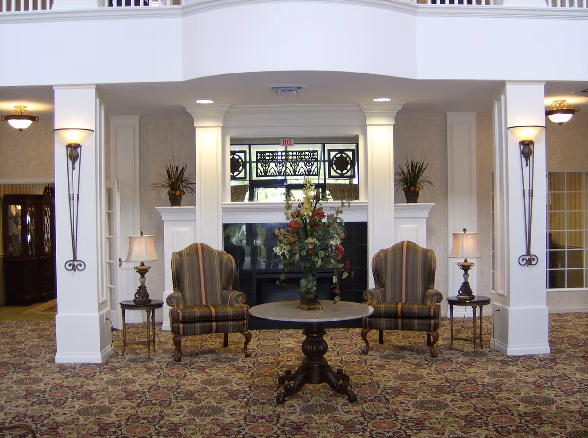 A cozy seating area in a senior living facility with two striped armchairs flanking a round wooden table that holds a floral arrangement. The area features a fireplace with decorative molding, two side tables with lamps, wall sconces, and patterned carpet. The space is well-lit and has a classic, elegant design.