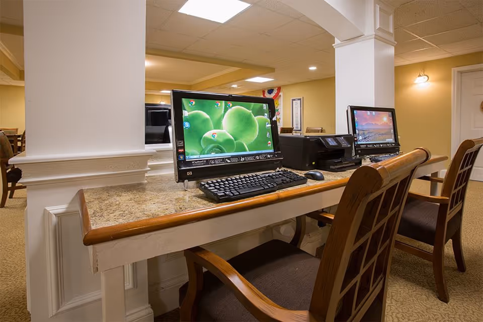 A communal computer workstation in a senior living facility showing two desktop computers, a printer, and chairs along a countertop in a carpeted common area.
