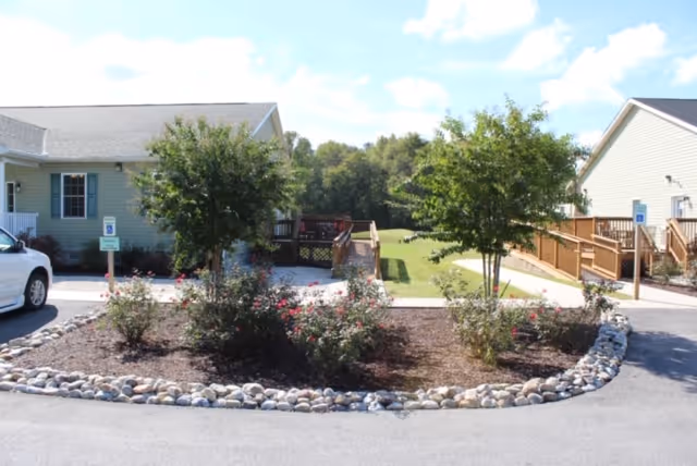 View of a landscaped area with small trees and bushes surrounded by a border of rocks, situated between two single-story buildings with ramps and parking spaces, under a partly cloudy sky.
