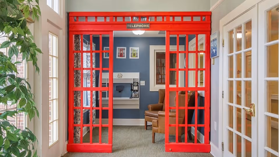 Interior view of a room with a decorative red British-style telephone booth frame. Inside the booth area, there is a vintage telephone mounted on the wall, a brown leather armchair, and framed pictures on a blue wall. To the left, there are tall windows with plants nearby, and to the right, a door with glass panels.