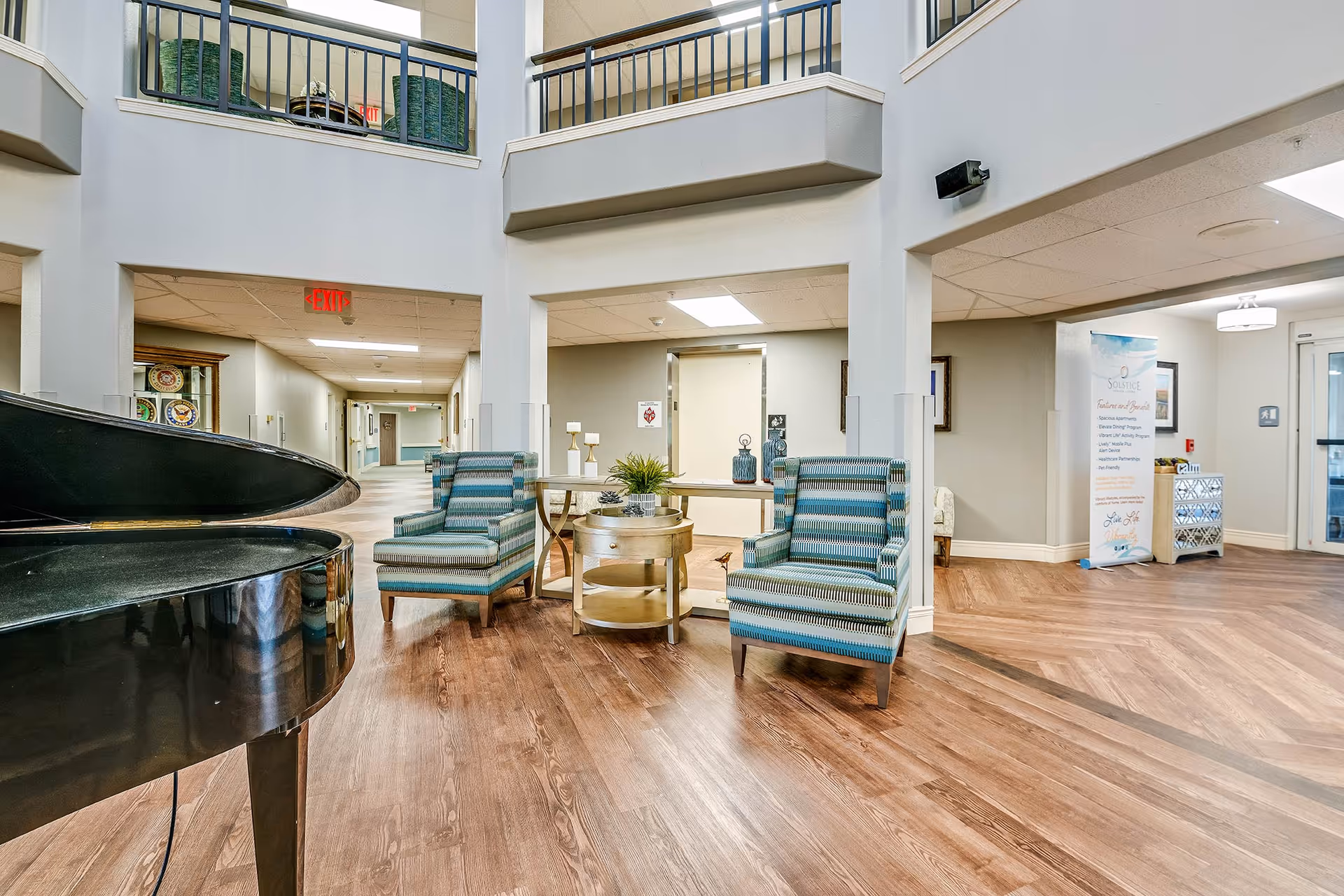 Interior view of a senior living facility lobby area with two striped armchairs and a round wooden side table between them. A black grand piano is partially visible on the left. The space has wood flooring, light-colored walls, and a high ceiling with a balcony railing above. There is an elevator in the background and a banner with the Solstice Senior Living logo and information on the right side near the entrance door.