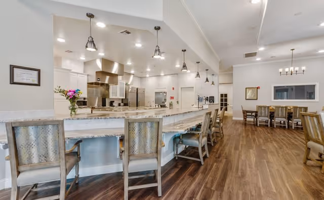 A spacious and modern kitchen and dining area in a senior living facility. The kitchen features stainless steel appliances, including a stove and refrigerator, with a long marble countertop bar lined with wooden chairs. Pendant lights hang above the bar. In the background, there are additional dining tables and chairs on a wooden floor, with neutral-colored walls and ceiling lights.