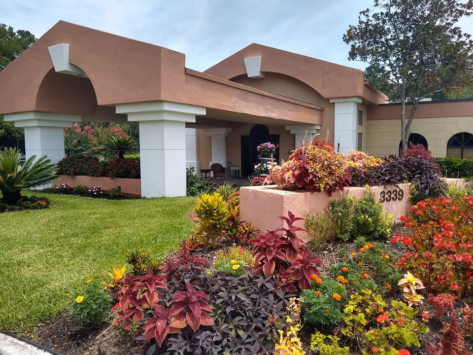 Exterior view of Diamond Assisted Living & Memory Care building entrance with a covered driveway, surrounded by well-maintained colorful flower beds and green grass under a partly cloudy sky.