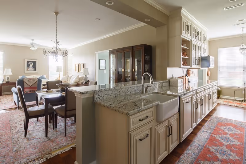 Bright and spacious open-concept living area featuring a kitchen with granite countertops, white cabinetry, and a farmhouse sink. Adjacent to the kitchen is a dining area with a wooden table and chairs, and a living room with sofas, armchairs, lamps, and framed artwork on the walls. Large windows allow natural light to fill the space, and decorative rugs cover the hardwood floors.