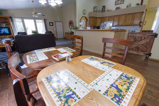 Communal dining area with wooden tables and sunflower placemats in front of an open kitchen and living room seating.